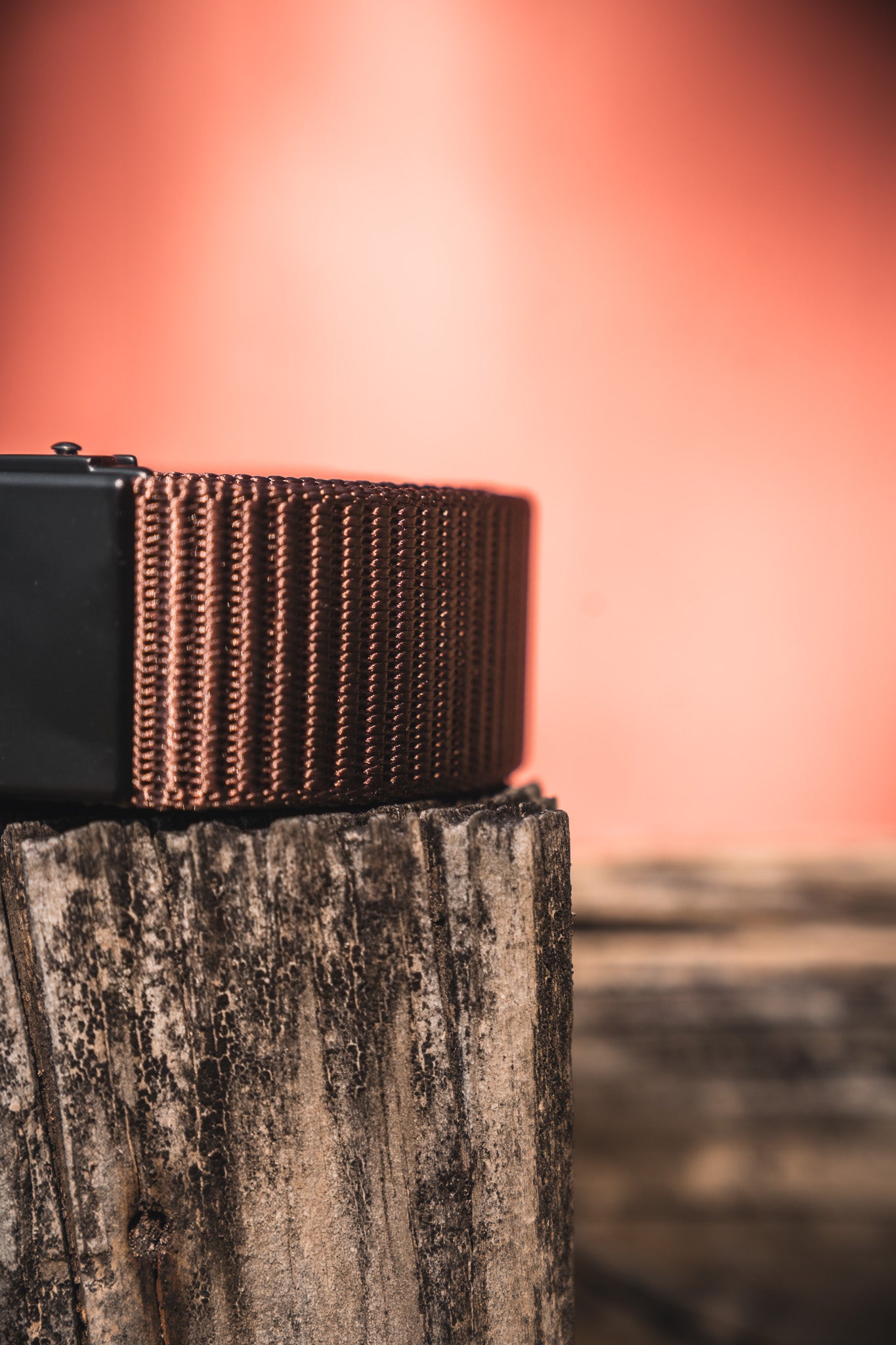 Close-up of the BU Logo Belt - Brown by Born United Private Label, featuring an adjustable design, resting on a weathered wooden surface with a blurred peach-colored background.