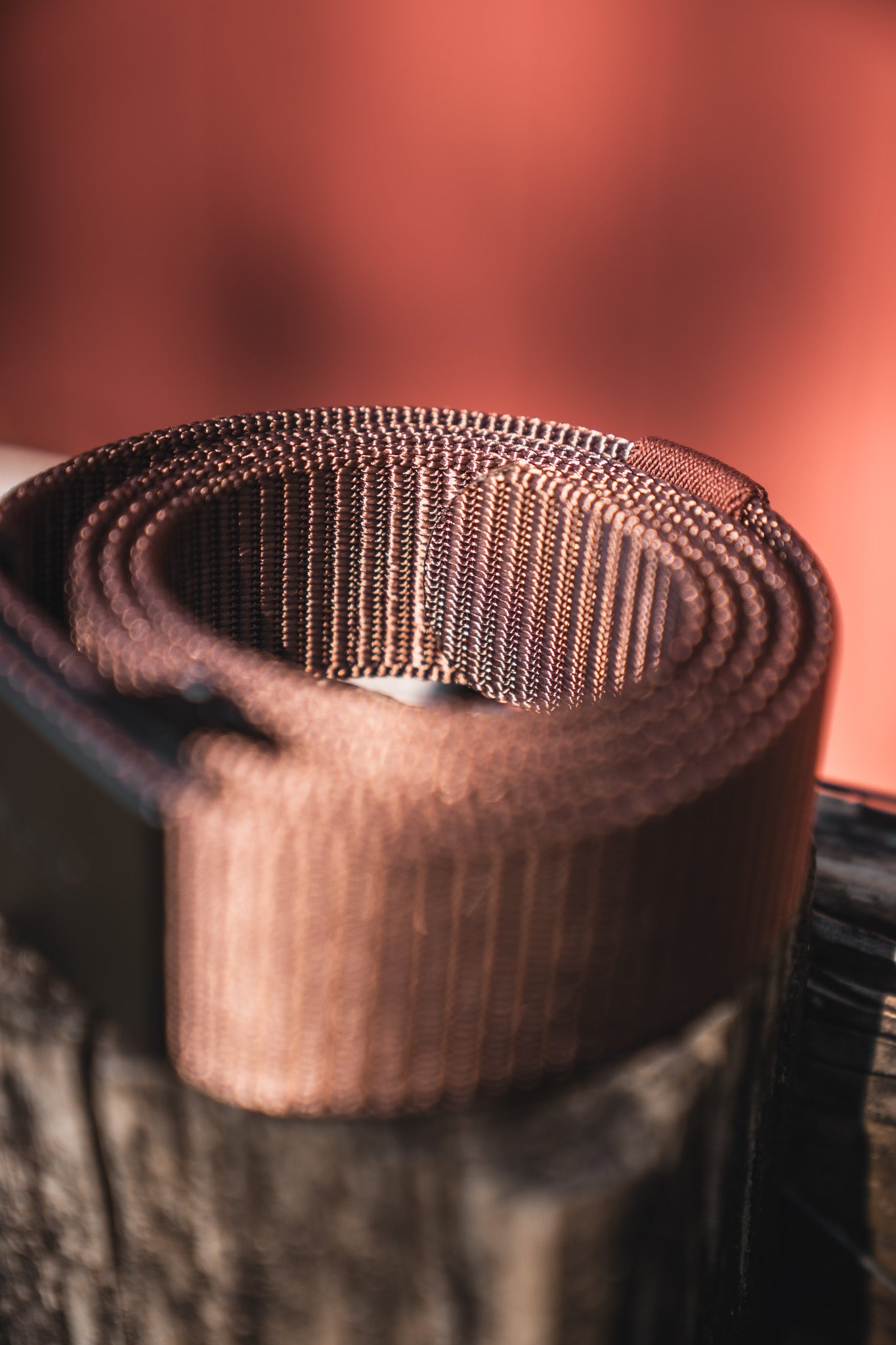 A close-up of the BU Logo Belt - Brown by Born United Private Label, an adjustable men's brown nylon belt, rolled up on a wooden surface with a blurred reddish background.