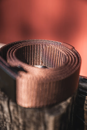 A close-up of the BU Logo Belt - Brown by Born United Private Label, an adjustable men's brown nylon belt, rolled up on a wooden surface with a blurred reddish background.