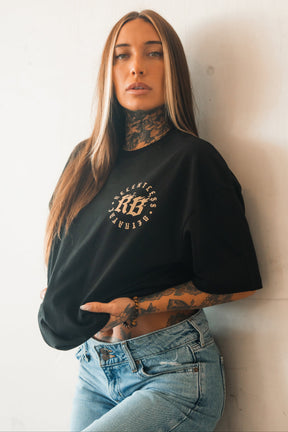 A woman with long, straight hair and visible tattoos poses against a white wall wearing the Relentless Betrayal Never Surrender Premium Oversized Ghost T-Shirt and light blue jeans, her hand resting confidently on her waist.