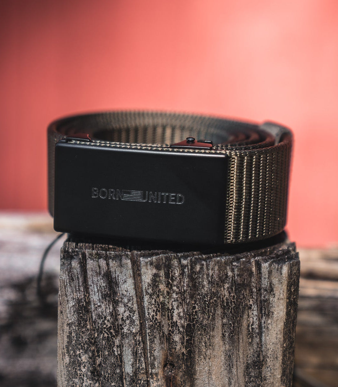 The BU Logo Belt - Army Green by Born United Private Label, featuring an adjustable design and a rectangular buckle engraved with BORN UNITED, is coiled on a weathered wooden post against a blurred reddish backdrop.