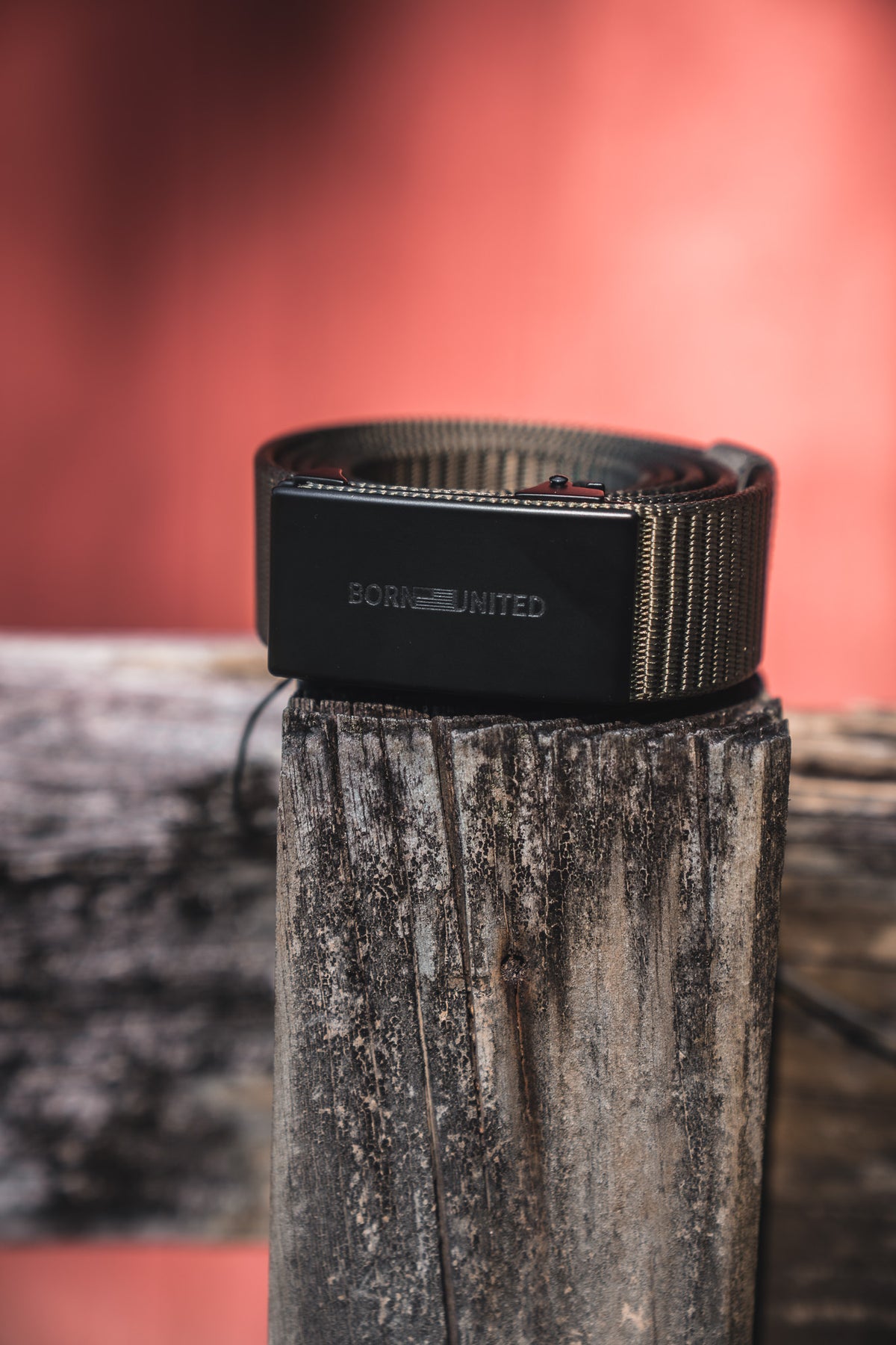 The BU Logo Belt - Army Green by Born United Private Label, featuring an adjustable design and a rectangular buckle engraved with BORN UNITED, is coiled on a weathered wooden post against a blurred reddish backdrop.