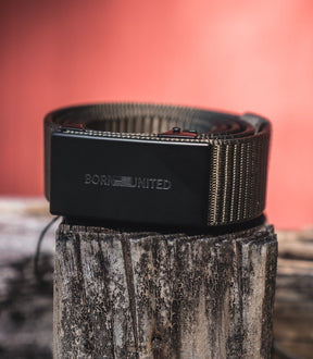 The BU Logo Belt - Army Green by Born United Private Label, featuring an adjustable design and a rectangular buckle engraved with BORN UNITED, is coiled on a weathered wooden post against a blurred reddish backdrop.