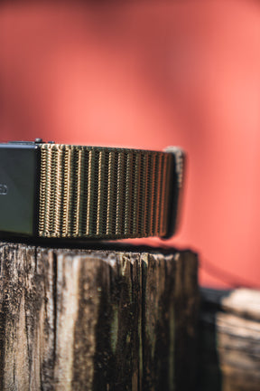 A close-up of the BU Logo Belt in army green by Born United Private Label, resembling an adjustable strap, displayed on a weathered wooden post with a blurred reddish background.