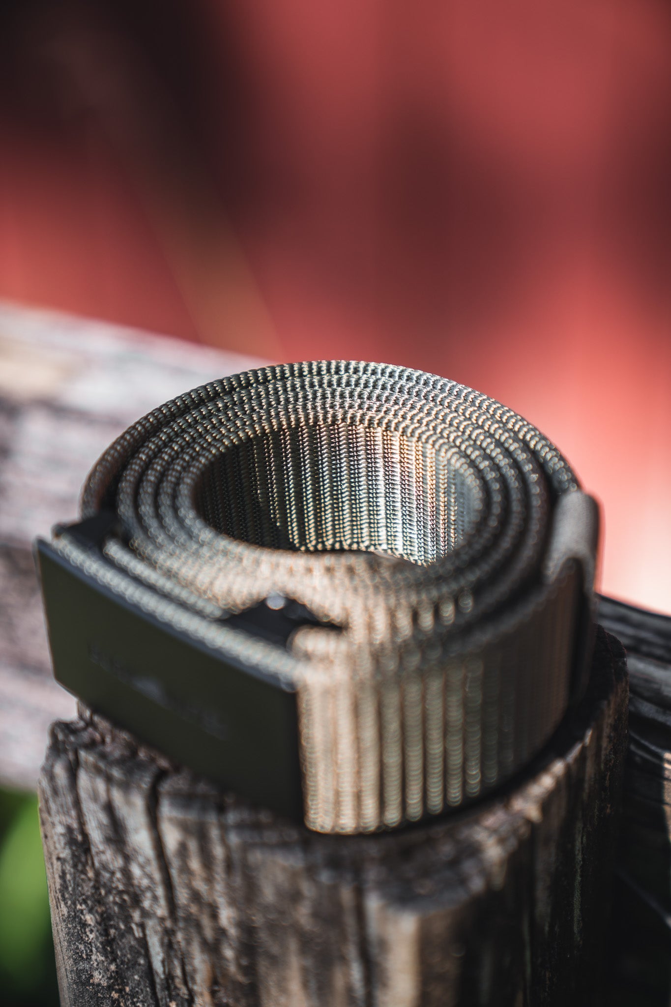 A close-up of the BU Logo Belt - Army Green by Born United Private Label, coiled with a textured finish and black buckle on a weathered wooden post. The blurred background features shades of red and brown.