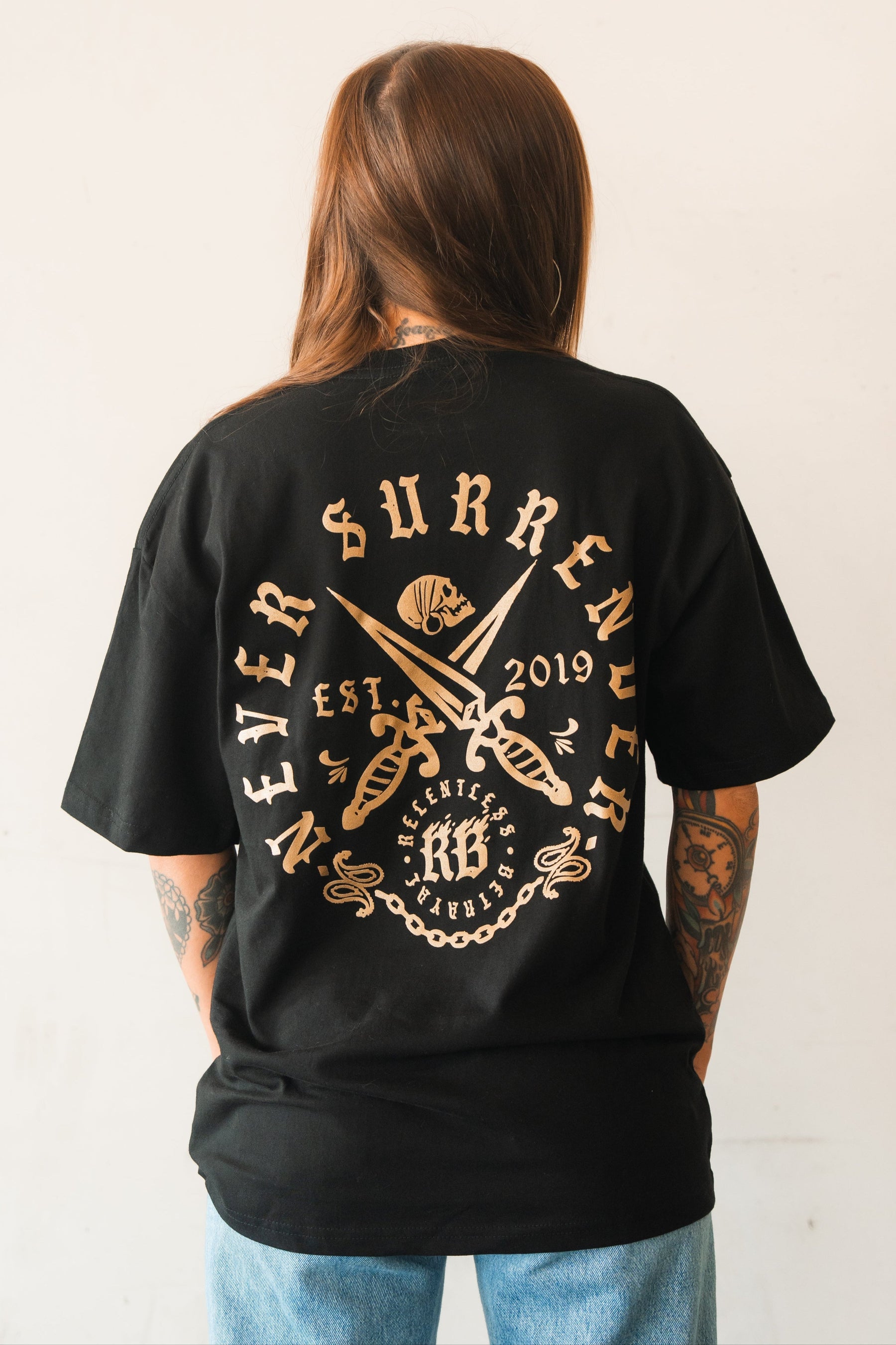 A person with long brown hair and tattoos wears the Relentless Betrayal Never Surrender Premium Oversized Ghost T-Shirt, featuring a bold back graphic of crossed swords, a skull, “Never Surrender,” “Est. 2019,” and decorative text.