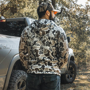 A man in a Mucker Bucket Wood Ducks Premium Athletic Duck Camo Hoodie and cap stands by a silver truck in a sunlit, wooded area, showcasing the hoodie’s quality in a natural outdoor setting.