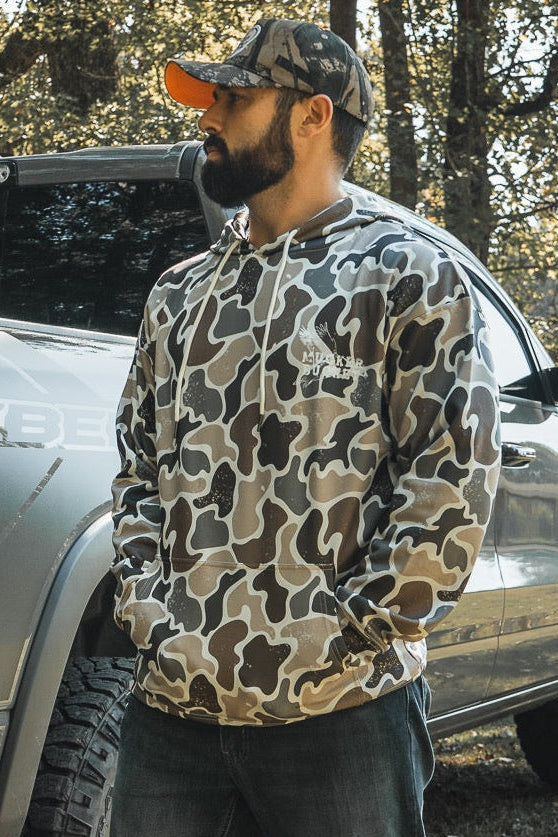 A bearded man in a Mucker Bucket Wood Ducks Premium Athletic Duck Camo Hoodie and cap stands in front of a silver pickup outdoors, with trees and sunlight in the background.