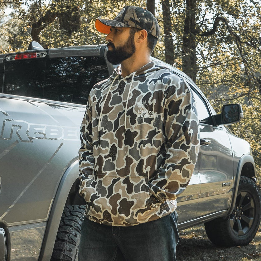 A bearded man in a Mucker Bucket Wood Ducks Premium Athletic Duck Camo Hoodie and cap stands in front of a silver pickup outdoors, with trees and sunlight in the background.
