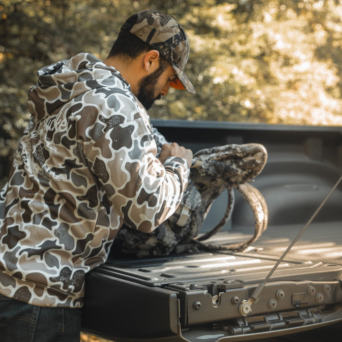A man in a Mucker Bucket Wood Ducks Premium Athletic Duck Camo Hoodie and cap stands at a pickup truck's open tailgate, looking into a camo backpack as sunlight filters through the trees in the background.