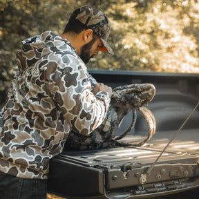 A man in a Mucker Bucket Wood Ducks Premium Athletic Duck Camo Hoodie and cap stands at a pickup truck's open tailgate, looking into a camo backpack as sunlight filters through the trees in the background.