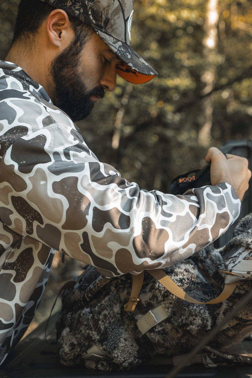 A man in a Mucker Bucket Wood Ducks Premium Athletic Duck Camo Hoodie and cap loads gear into a camo backpack on the open tailgate of a truck in a wooded outdoor setting.