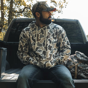 A bearded man in a Mucker Bucket Wood Ducks Premium Athletic Duck Camo Hoodie and cap sits on a pickup tailgate in sunlight, with a camo-patterned bag by his side and trees in the background.