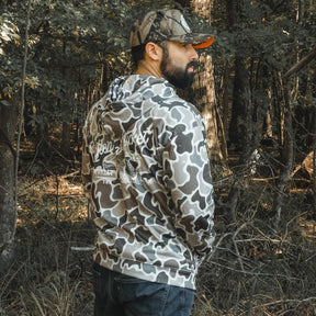 A bearded man stands in a forest, wearing a Mucker Bucket Wood Ducks Premium Athletic Duck Camo Hoodie and a matching camo cap, looking to the side with his back partially facing the camera amid trees and foliage in the background.