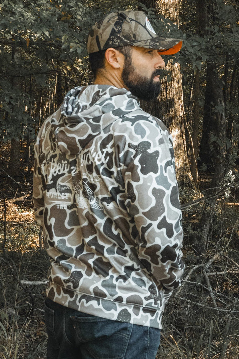A bearded man stands in a forest, wearing a Mucker Bucket Wood Ducks Premium Athletic Duck Camo Hoodie and a matching camo cap, looking to the side with his back partially facing the camera amid trees and foliage in the background.