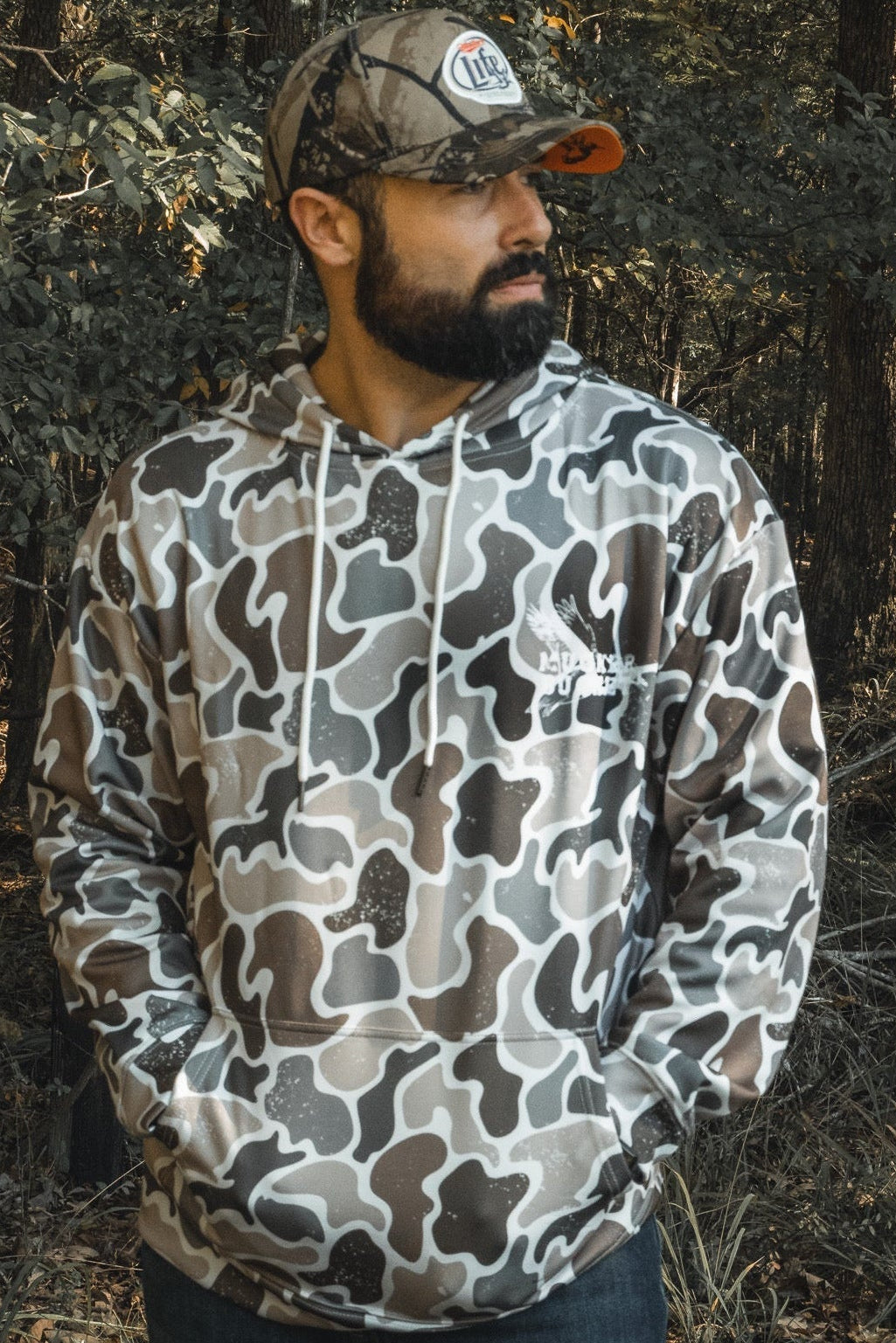 A bearded man stands in a forest with hands in his pockets, wearing a Mucker Bucket Wood Ducks Premium Athletic Duck Camo Hoodie and matching hat, sunlight streaming through the trees behind him.