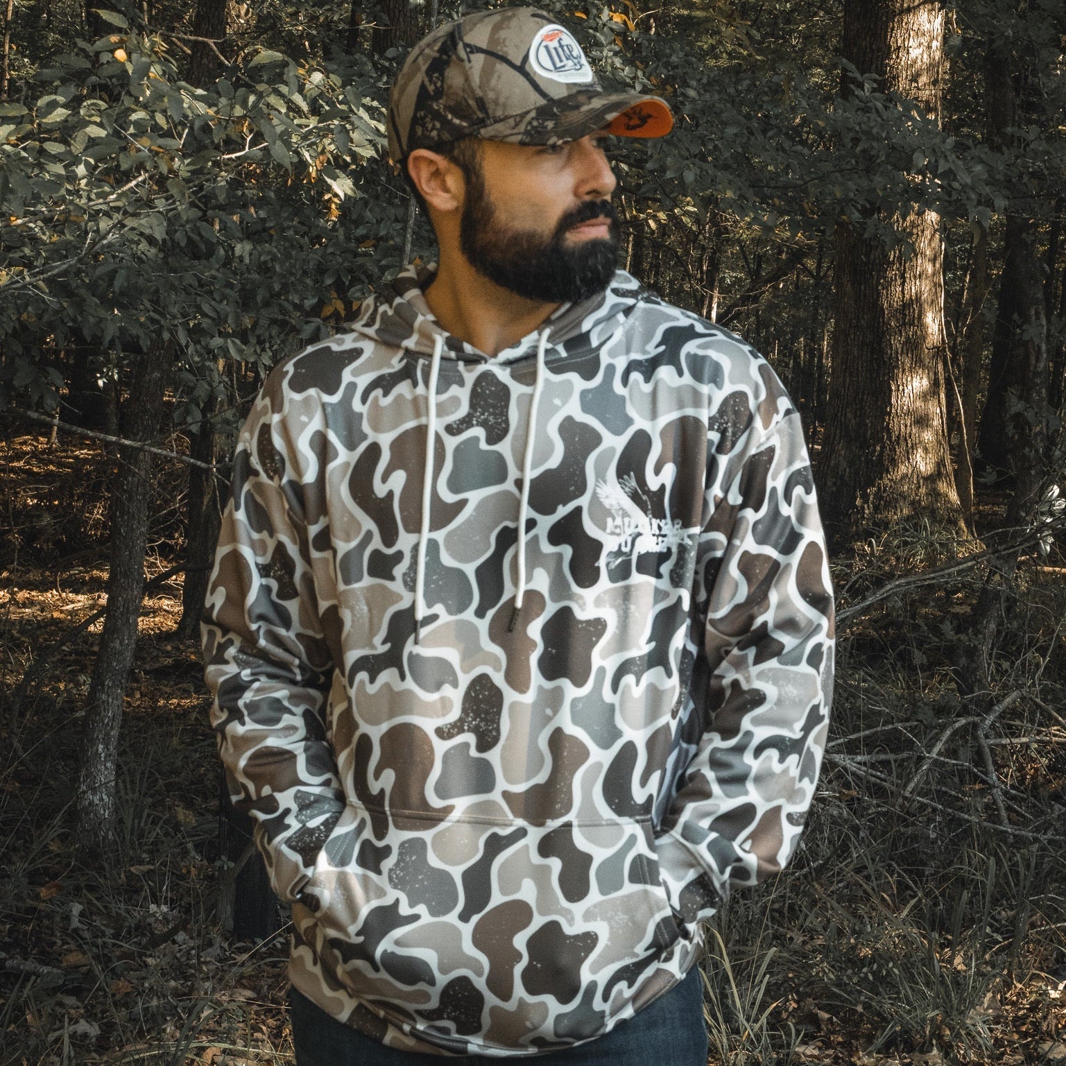 A bearded man stands in a forest with hands in his pockets, wearing a Mucker Bucket Wood Ducks Premium Athletic Duck Camo Hoodie and matching hat, sunlight streaming through the trees behind him.