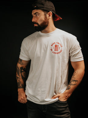 A bearded man, wearing the Legends - Shrimp Co. white graphic t-shirt and a black cap from Born United Private Label, stands against a dim backdrop. His tattooed arms and serious leftward gaze create an enigmatic presence.