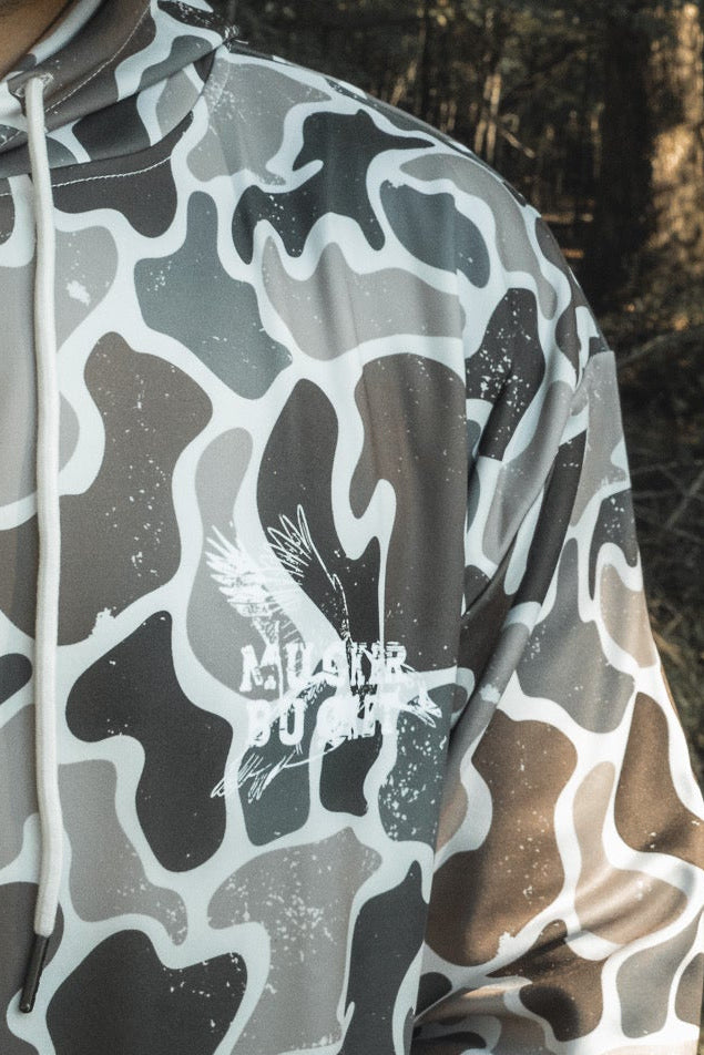 A person wears a gray and white Wood Ducks Premium Athletic Duck Camo Hoodie by Mucker Bucket, featuring a bird graphic and "ALDER POWER" text, standing outdoors in a wooded area.