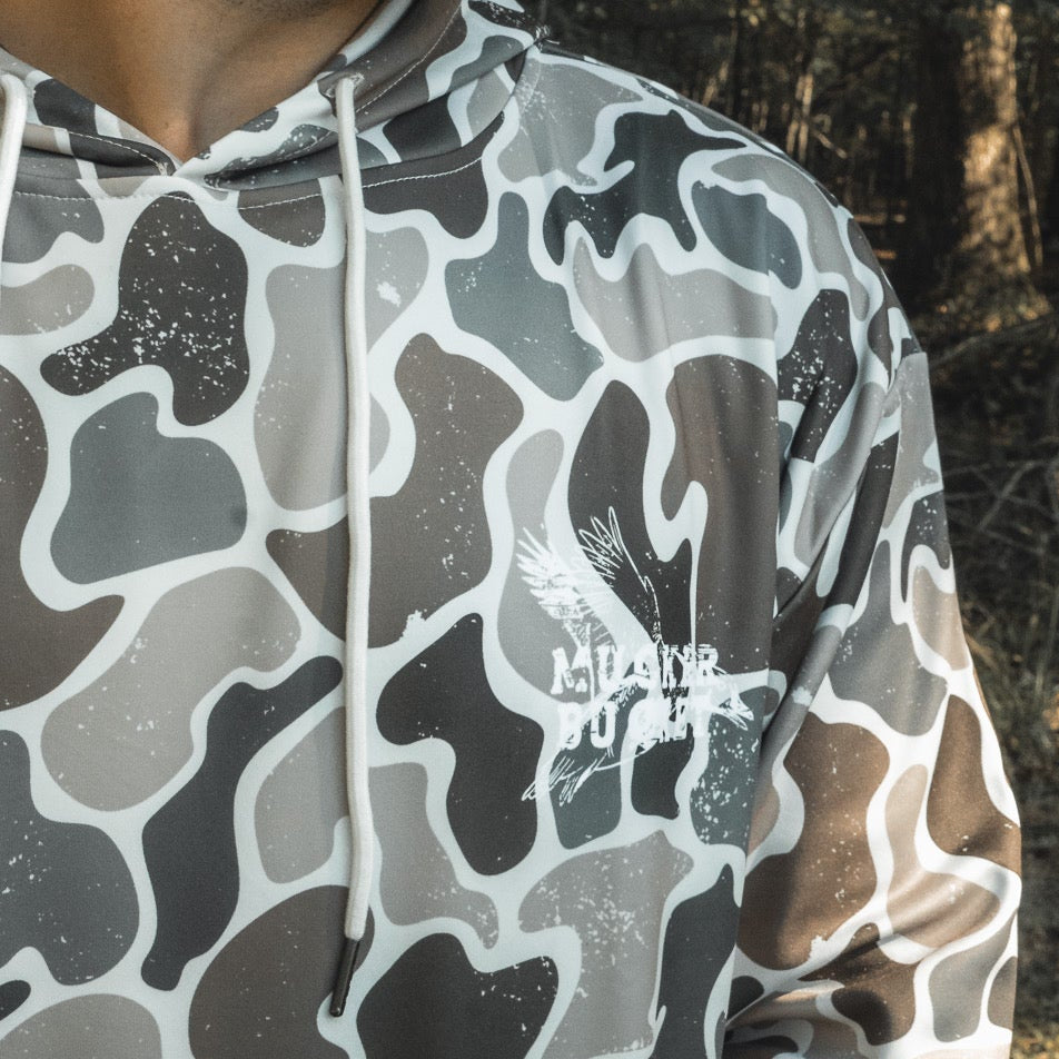 A person wears a gray and white Wood Ducks Premium Athletic Duck Camo Hoodie by Mucker Bucket, featuring a bird graphic and "ALDER POWER" text, standing outdoors in a wooded area.