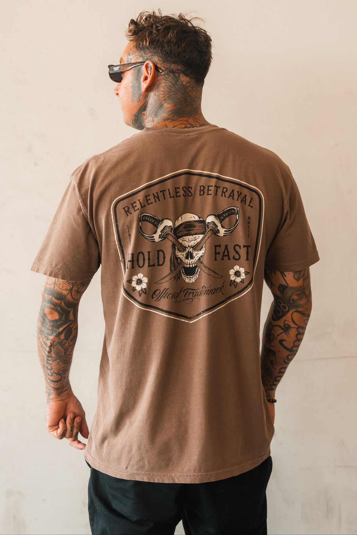 A tattooed man in sunglasses faces a light wall, wearing the Relentless Betrayal Hold Fast Premium Cocoa T-Shirt made from heavyweight jersey cotton. The shirt features a graphic snake and bold HOLD FAST print for lasting comfort and style.