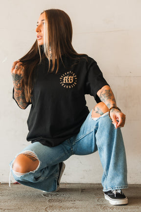 A woman with long hair and tattoos poses against a light wall, crouching in ripped blue jeans and sneakers, wearing the Relentless Betrayal Never Surrender Premium Oversized Ghost T-Shirt with a circular design.