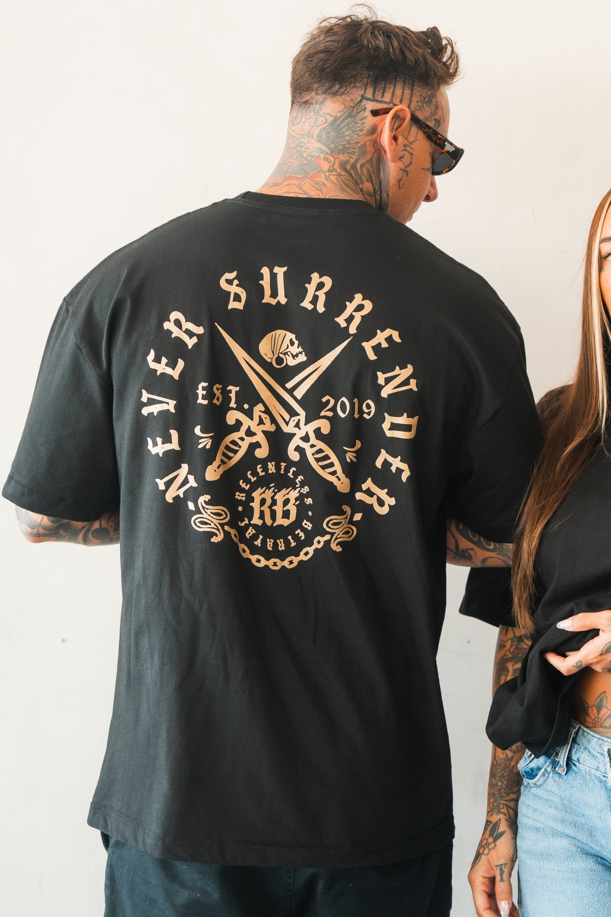 A tattooed man in sunglasses wears the Relentless Betrayal Never Surrender Premium Oversized Ghost T-Shirt, featuring a back graphic with "Never Surrender," crossed swords, and "EST. 2019." Another person’s arm appears beside him.