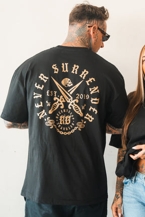 A tattooed man in sunglasses wears the Relentless Betrayal Never Surrender Premium Oversized Ghost T-Shirt, featuring a back graphic with "Never Surrender," crossed swords, and "EST. 2019." Another person’s arm appears beside him.