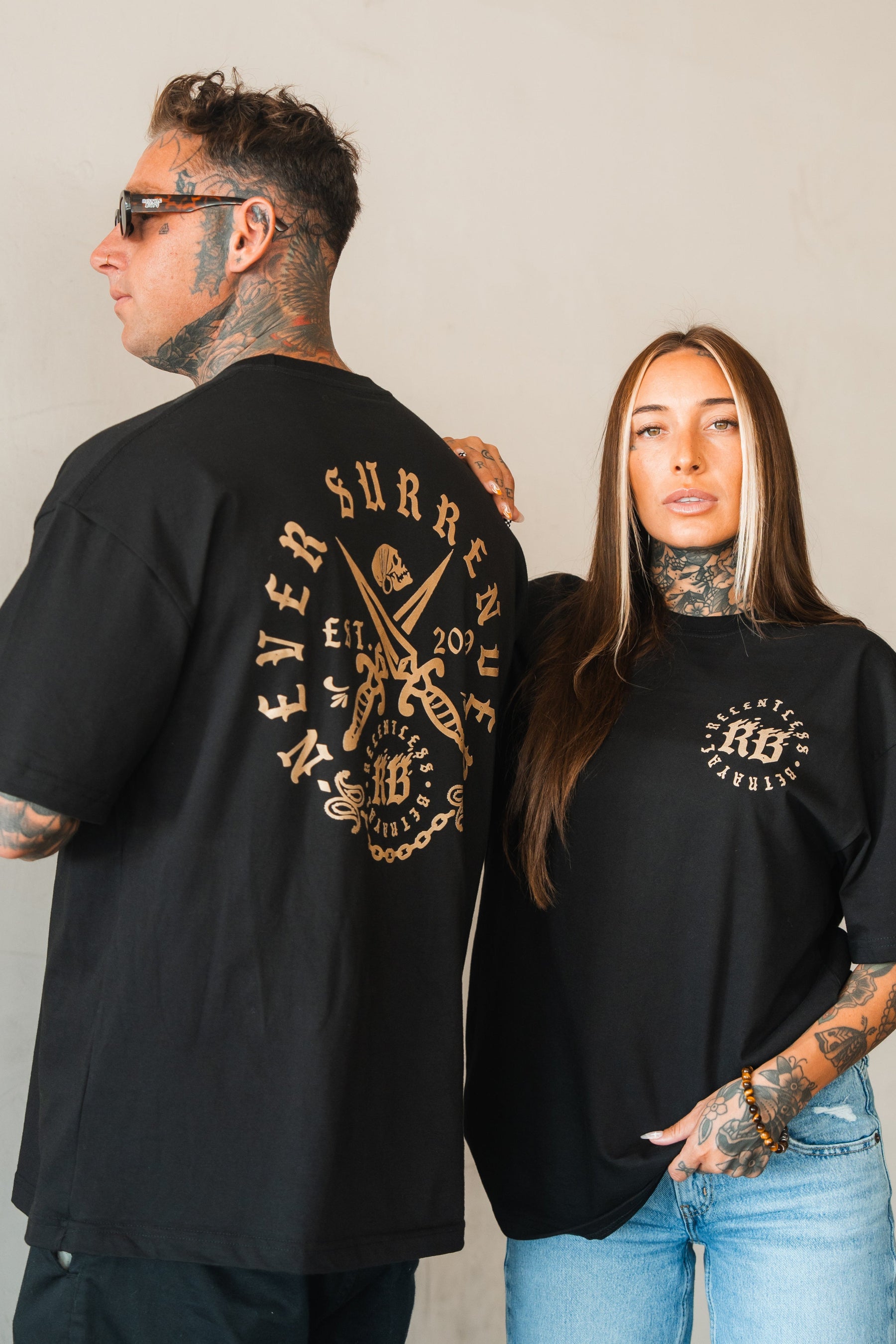 Two tattooed models with long hair wear Relentless Betrayal’s Never Surrender Premium Oversized Ghost T-Shirt: the man, seen from the side, shows the large back print, while the woman, facing forward, displays the chest logo.