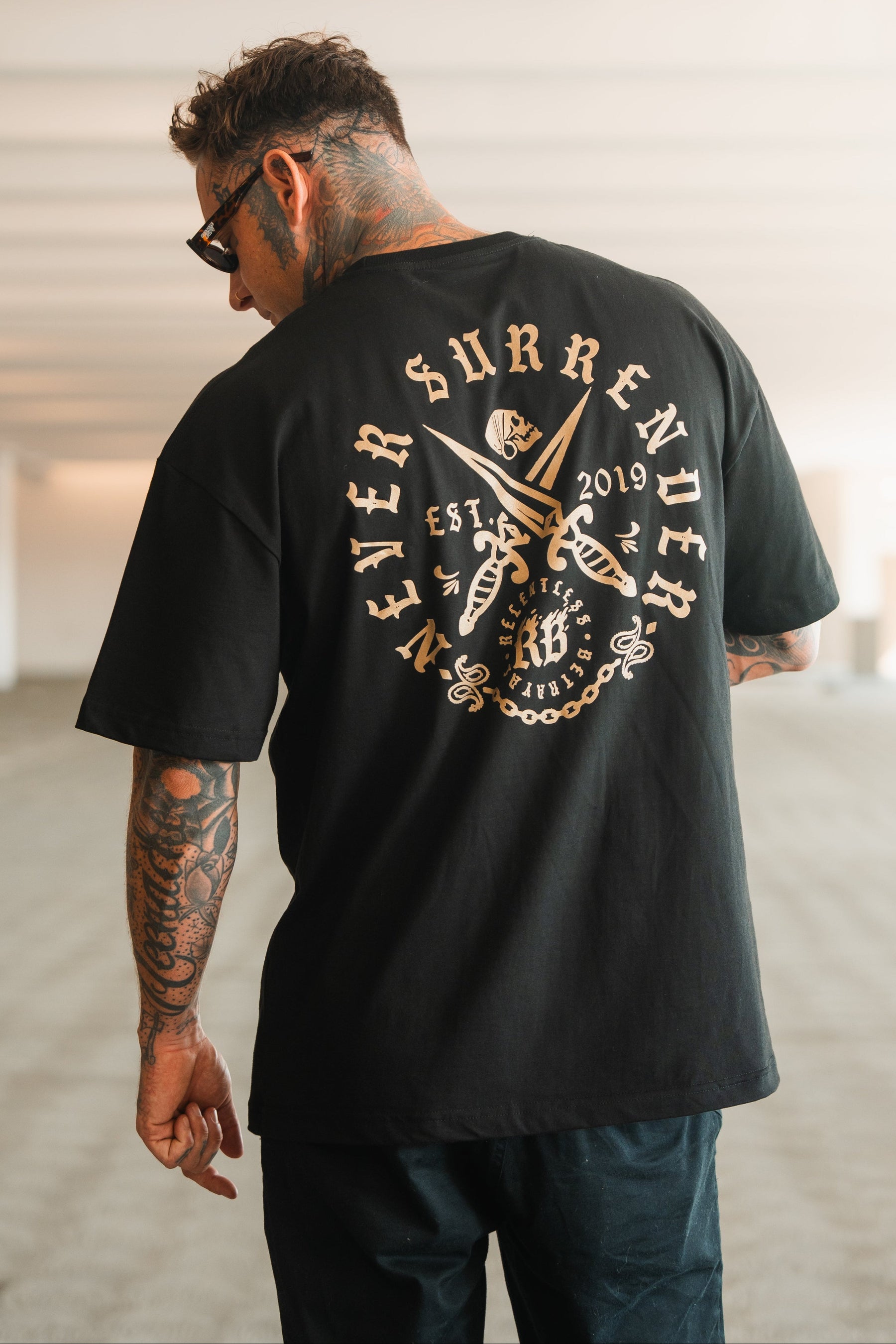 A tattooed man in sunglasses faces away, wearing the Relentless Betrayal Never Surrender Premium Oversized Ghost T-Shirt—a black heavyweight tee with crossed swords and bold gold “Never Surrender, EST 2019” lettering on the back.