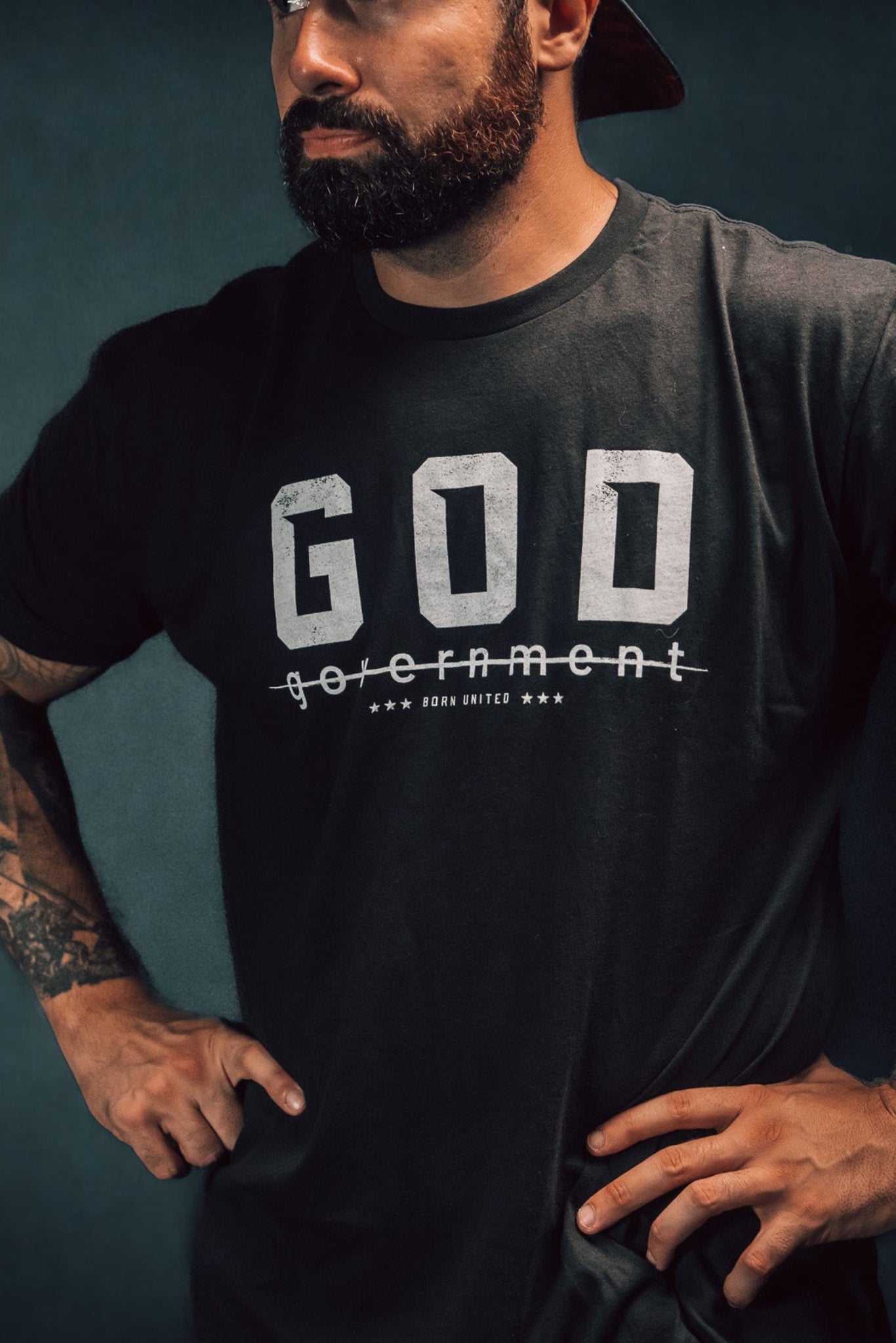 A bearded man, gazing to the side against a dark background, stands confidently with hands on hips. Hes wearing a reversed baseball cap and Born United Private Labels God Over Government t-shirt, which features the word government crossed out below GOD, challenging authority with his defiant posture.