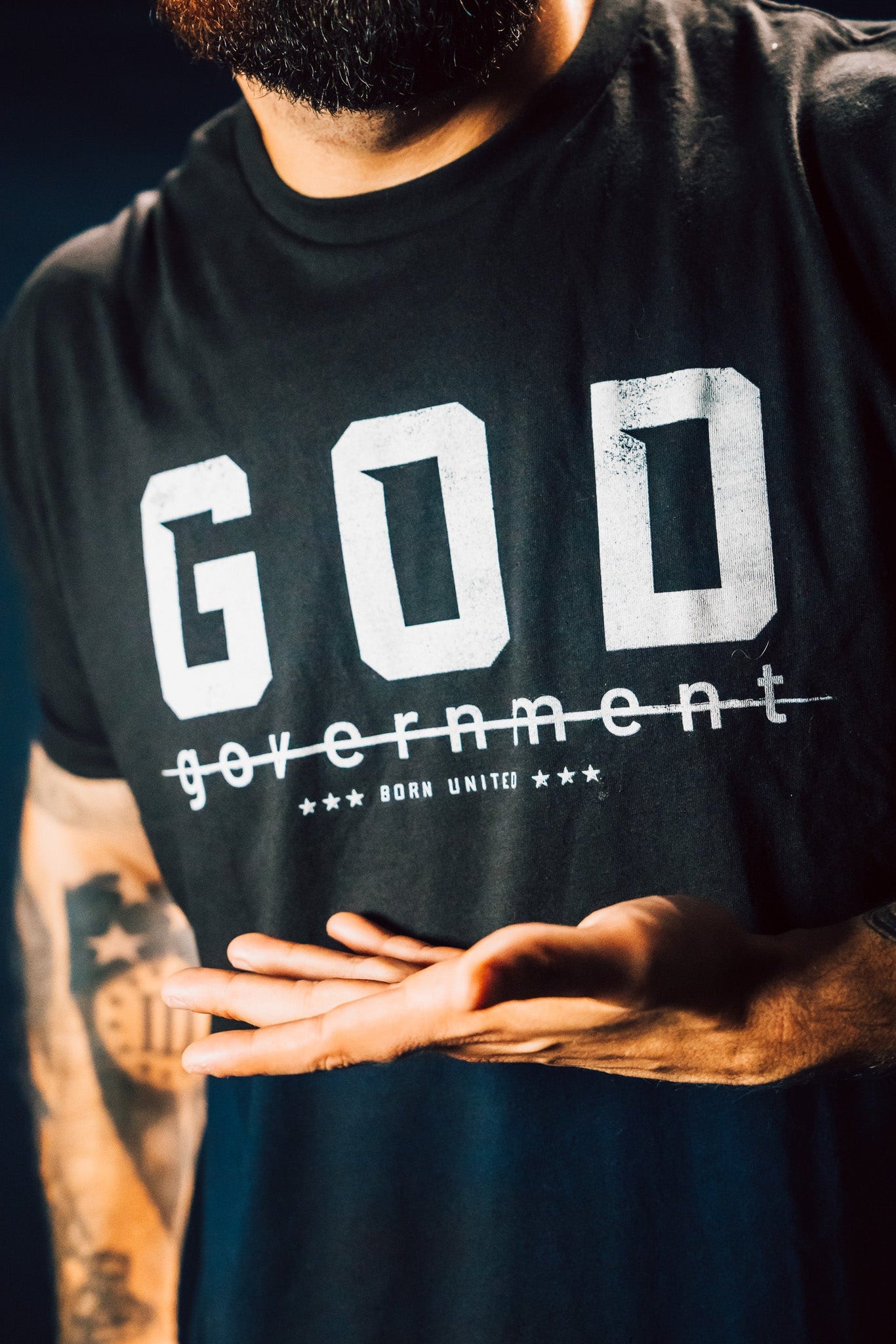 A person wearing a God Over Government black T-shirt by Born United Private Label, featuring stars and bold text, stands confidently in a dark background. Their tattooed arm gestures respect for rulers and good conduct.