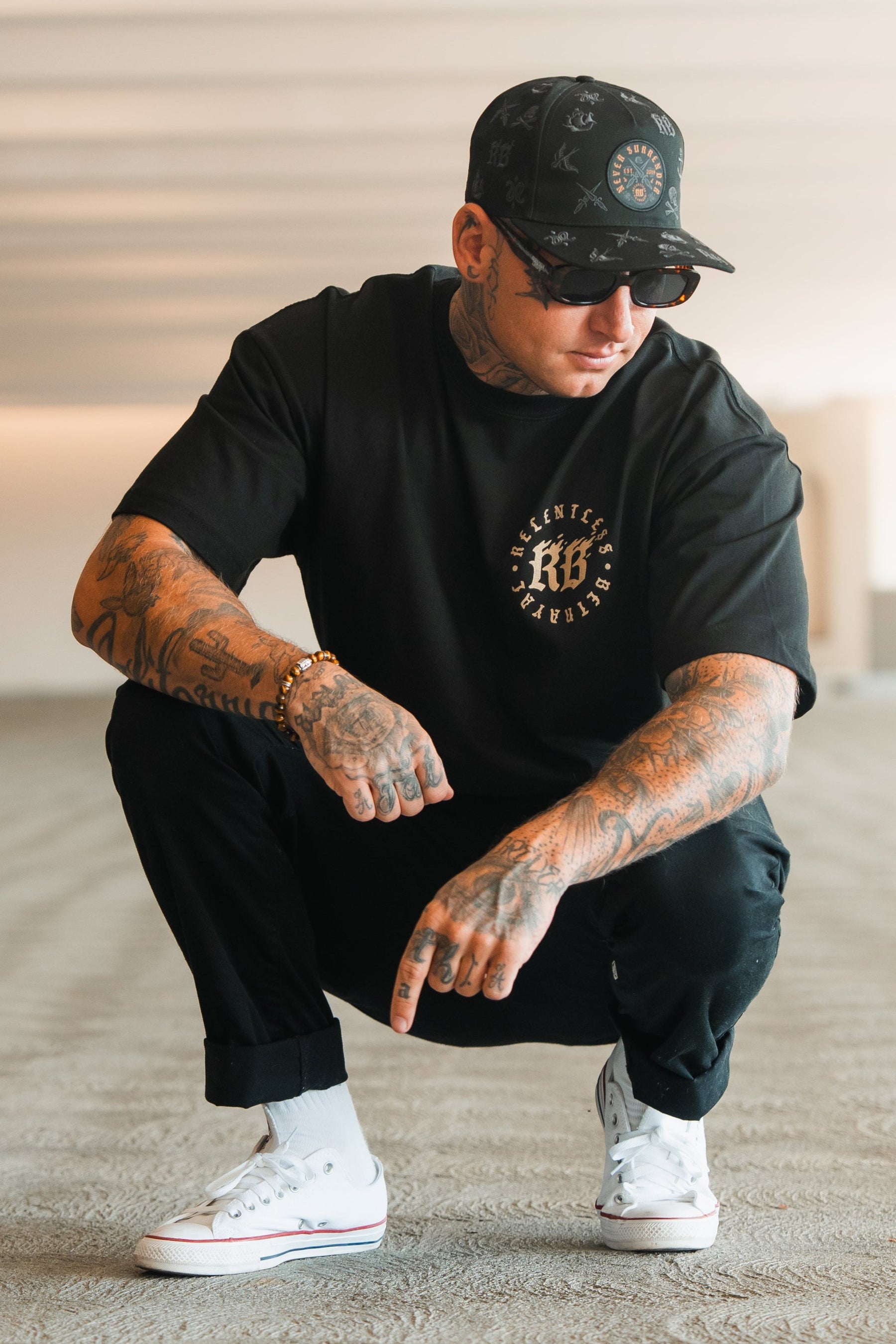 A tattooed man squats in a parking garage wearing Relentless Betrayal’s Never Surrender Premium Oversized Ghost T-Shirt, black pants, white Converse sneakers, a black cap, and sunglasses, looking down and pointing with one finger.
