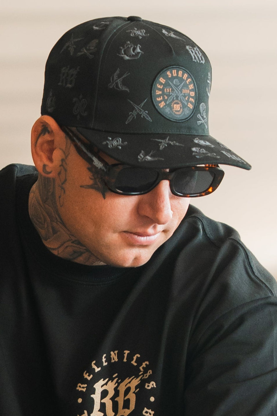 Wearing the Relentless Betrayal Never Surrender Limited Edition Snapback, black sunglasses, and a black Relentless shirt, a tattooed individual with visible face and neck ink looks downward with a neutral expression.