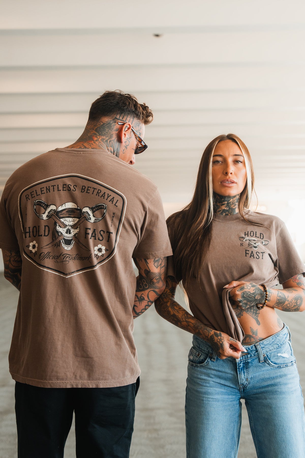 A tattooed man and woman wear Relentless Betrayal's Hold Fast Premium Cocoa T-Shirts in a bright, modern space. His features a skull, snakes, and "Relentless Betrayal, Hold Fast," while hers says "Hold Fast" on the front.