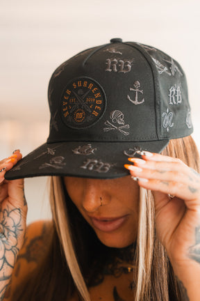 A person with long hair and tattooed hands holds the brim of a Relentless Betrayal Never Surrender Limited Edition Snapback, adorned with anchors, skulls, and bold lettering. Their nails are long, painted orange and white.
