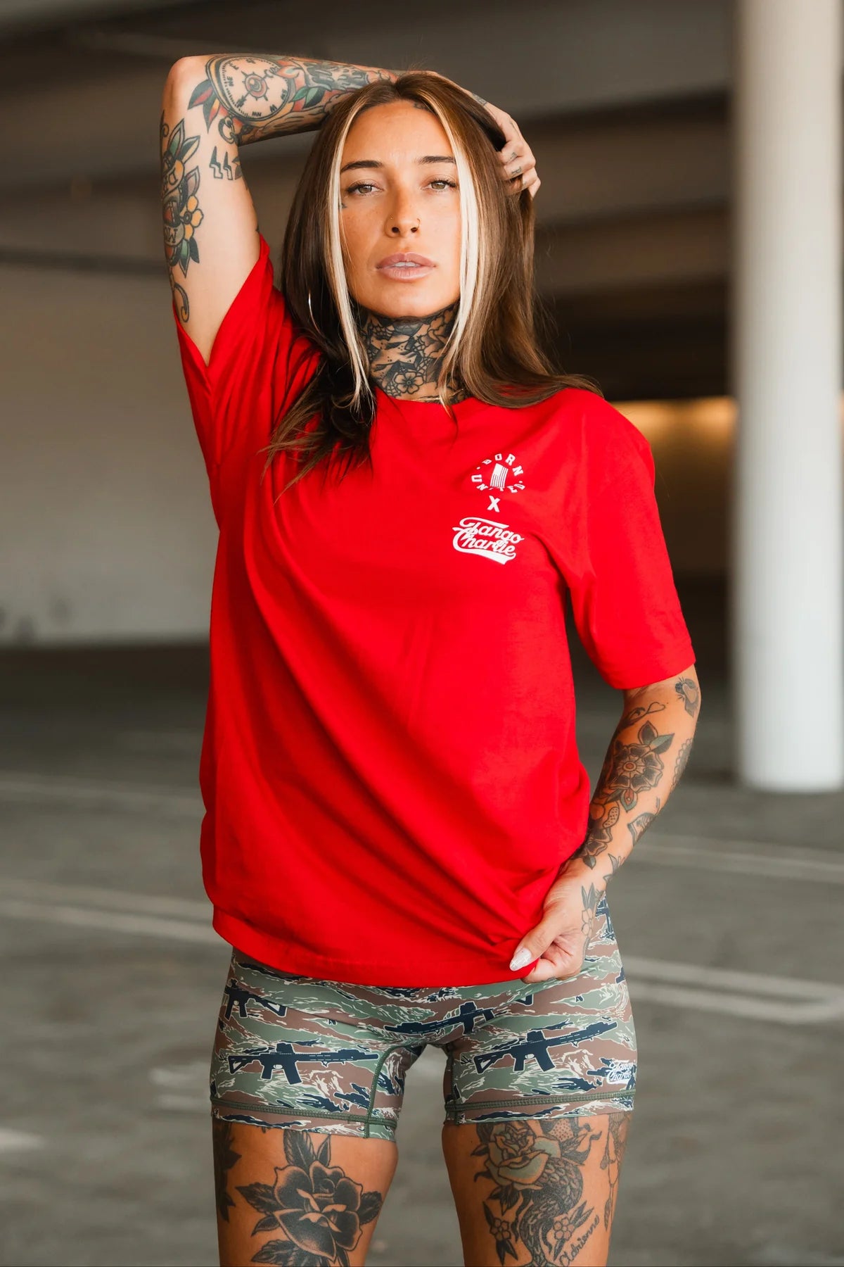 A tattooed woman poses confidently in a parking garage wearing the Tango Charlie Apparel “Make America Fit Again - Tee” and camouflage shorts, embodying health and wellness with one hand on her head and the other pulling her shirt.