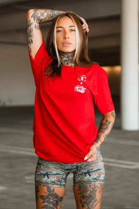 A tattooed woman poses confidently in a parking garage wearing the Tango Charlie Apparel “Make America Fit Again - Tee” and camouflage shorts, embodying health and wellness with one hand on her head and the other pulling her shirt.