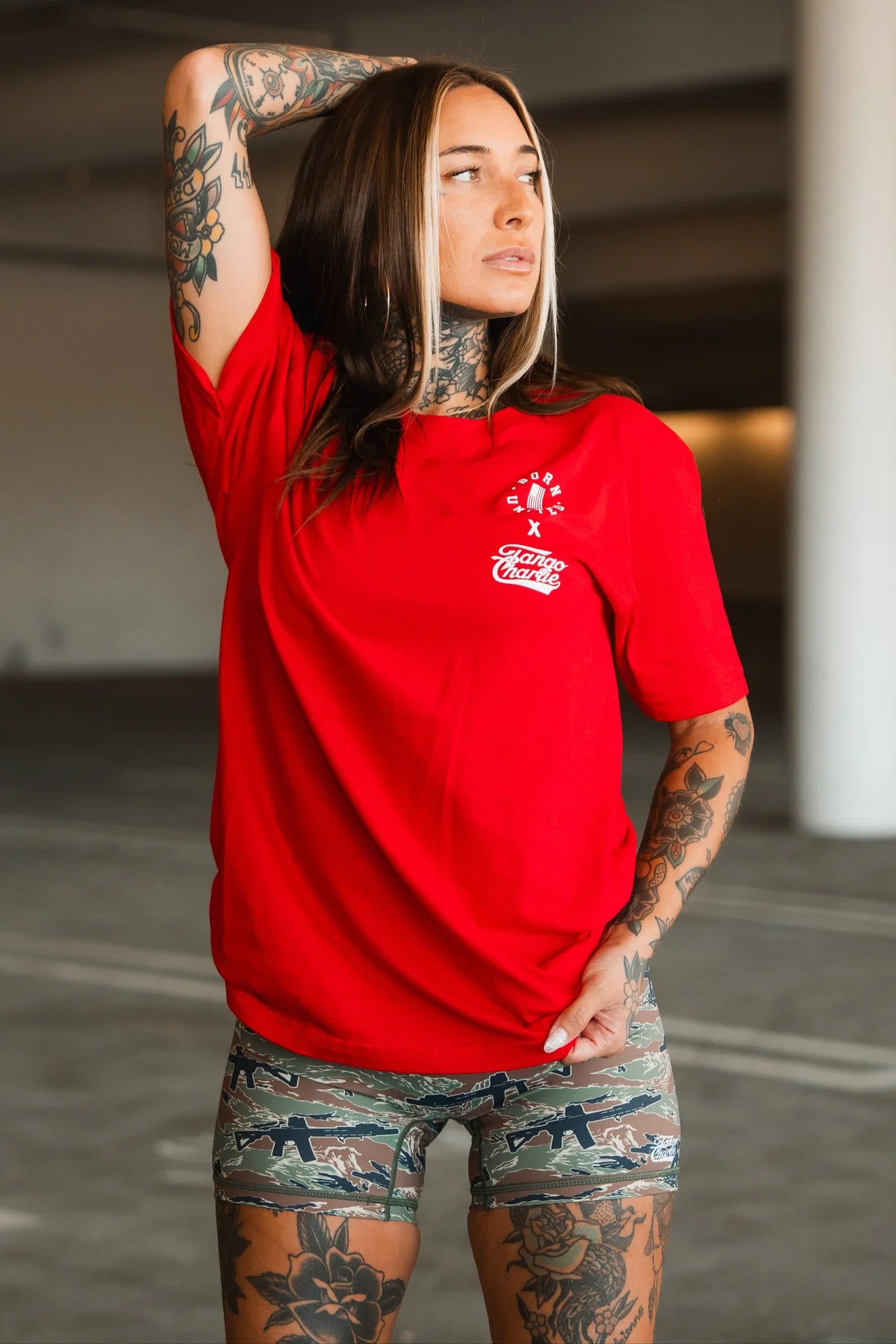 A tattooed woman models the Make America Fit Again Tee by Tango Charlie Apparel, pairing it with camo shorts as she stands confidently in an urban setting, capturing a bold and energetic approach to wellness.