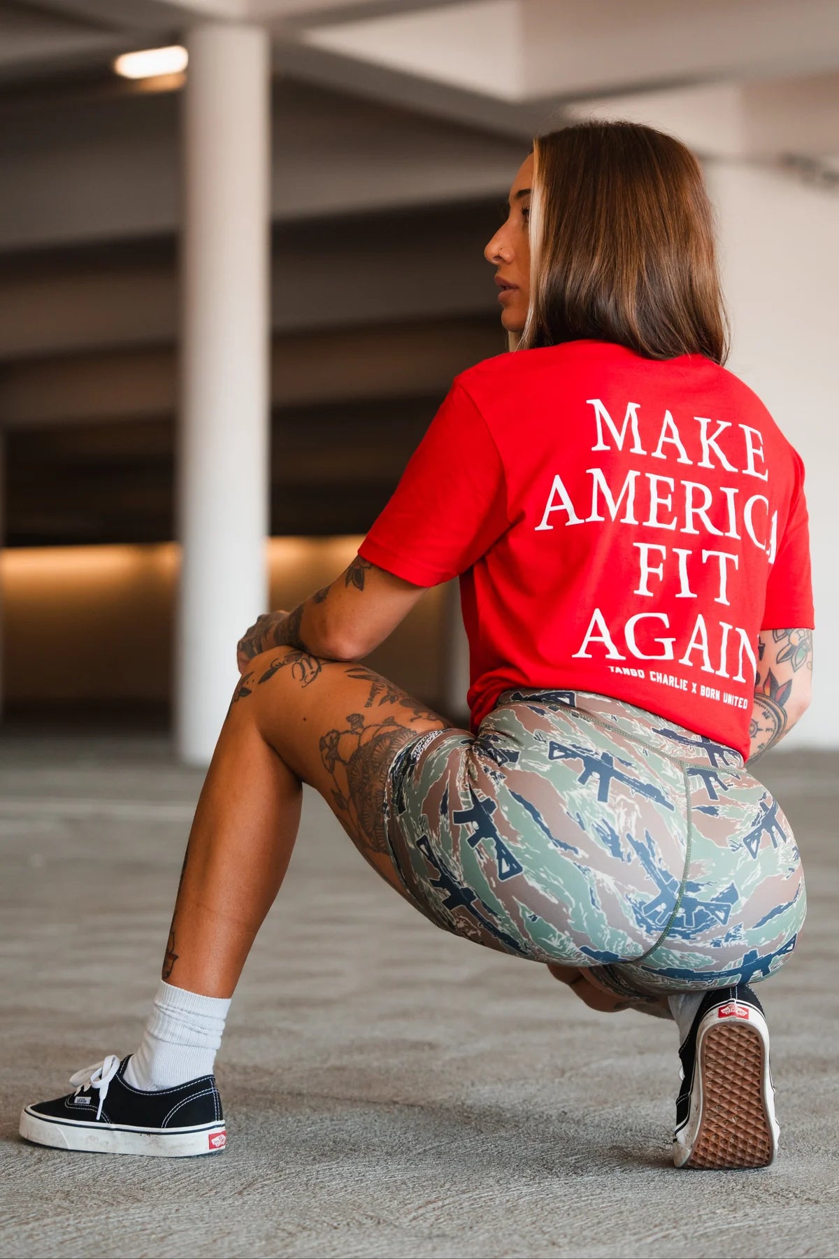 A health enthusiast with tattoos wears a red Make America Fit Again - Tee by Tango Charlie Apparel, paired with camouflage shorts, white socks, and black sneakers, while squatting indoors facing away from the camera.