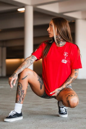 A tattooed person with long hair squats indoors, embracing wellness. They wear the bright red "Make America Fit Again - Tee" by Tango Charlie Apparel, black shorts, white socks, and black sneakers amid concrete pillars and soft lighting.