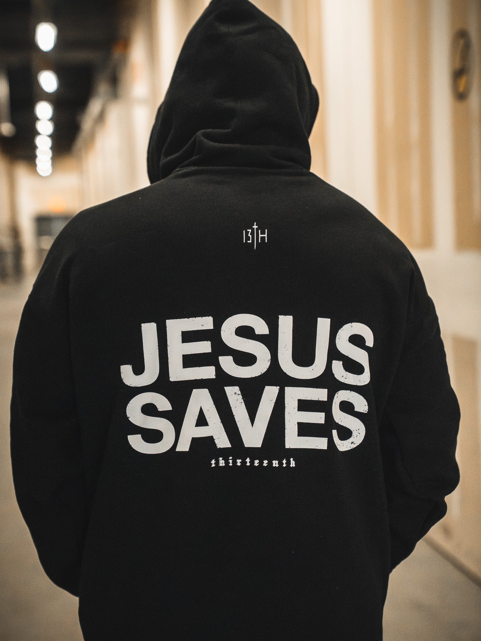 A person in a black 13th Jesus Saves Hoodie with bold white letters stands in a hallway; "Jesus Saves" is front and center, with "13TH" above and "shirtsernth" below, making a strong statement of Christian faith.