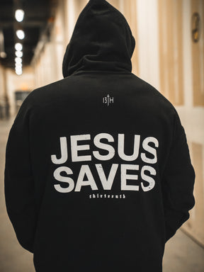 A person in a black 13th Jesus Saves Hoodie with bold white letters stands in a hallway; "Jesus Saves" is front and center, with "13TH" above and "shirtsernth" below, making a strong statement of Christian faith.
