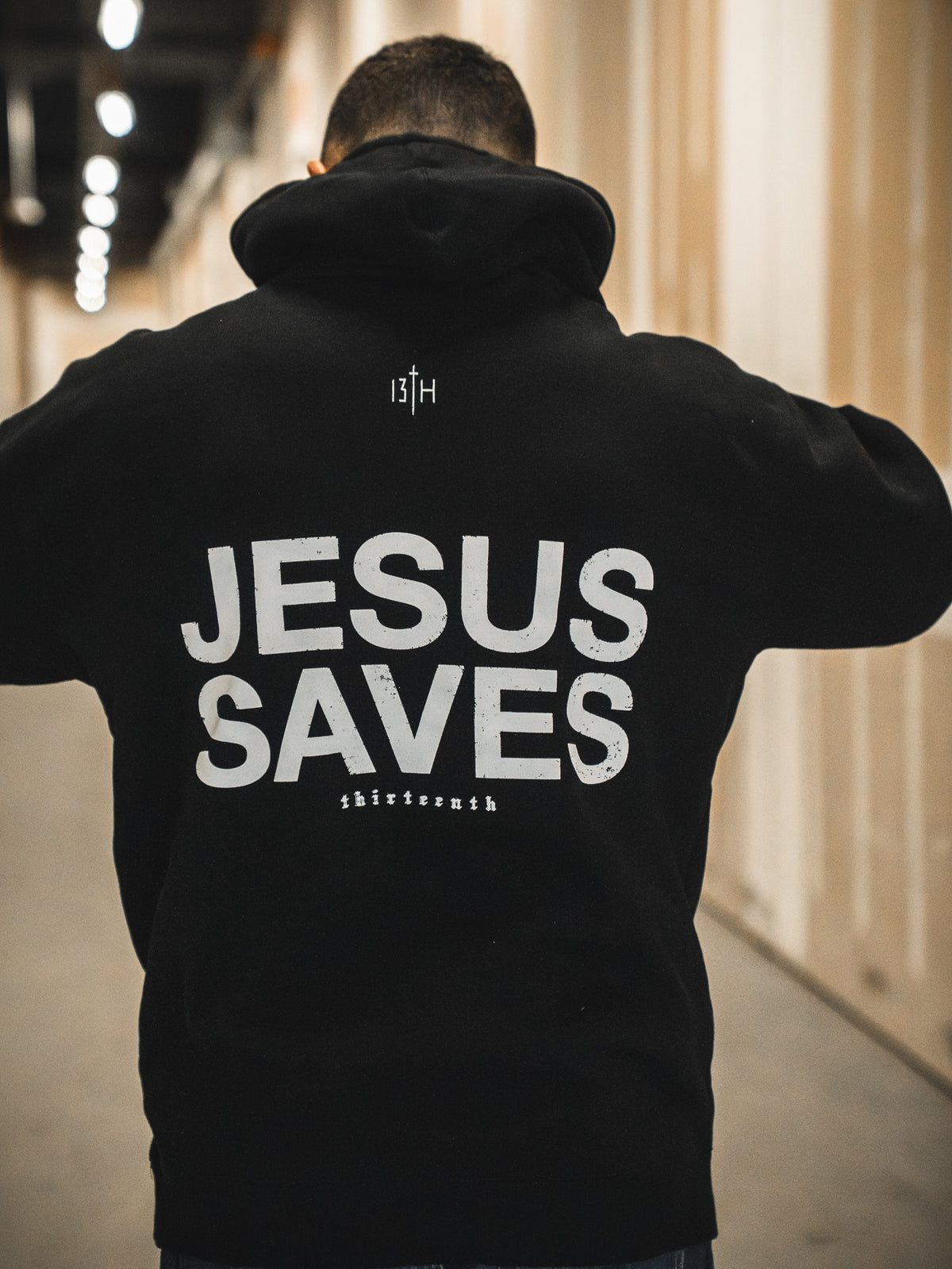 A person stands in a hallway wearing the 13th Jesus Saves Hoodie, featuring JESUS SAVES boldly printed on the back and "thirteenth" in smaller text near the bottom.