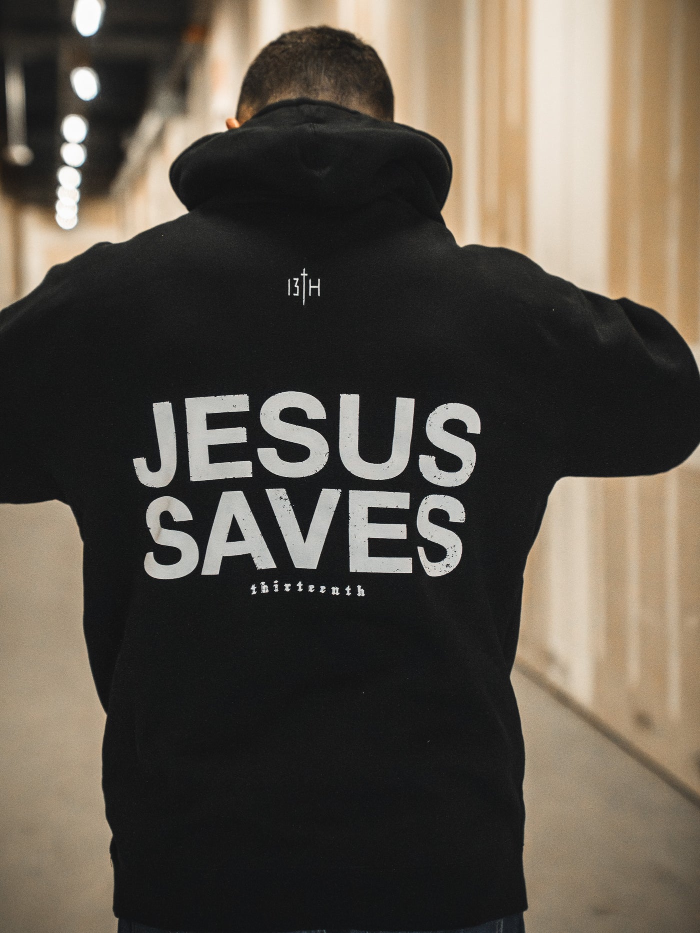 A person stands in a hallway wearing the 13th Jesus Saves Hoodie, featuring JESUS SAVES boldly printed on the back and "thirteenth" in smaller text near the bottom.