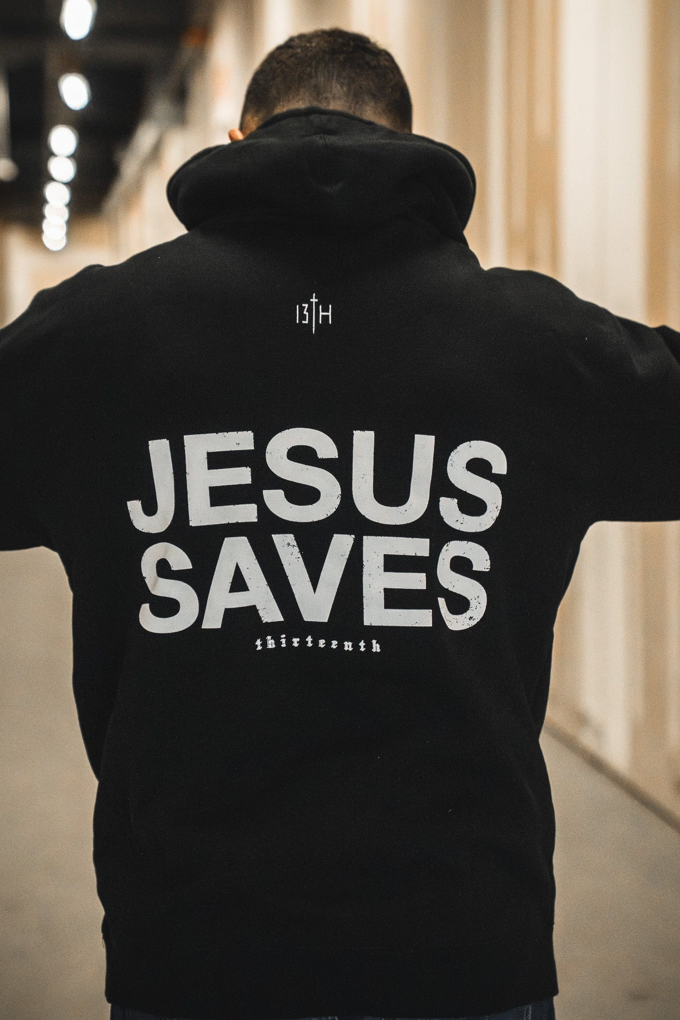 A person stands in a hallway wearing the 13th Jesus Saves Hoodie, featuring JESUS SAVES boldly printed on the back and "thirteenth" in smaller text near the bottom.