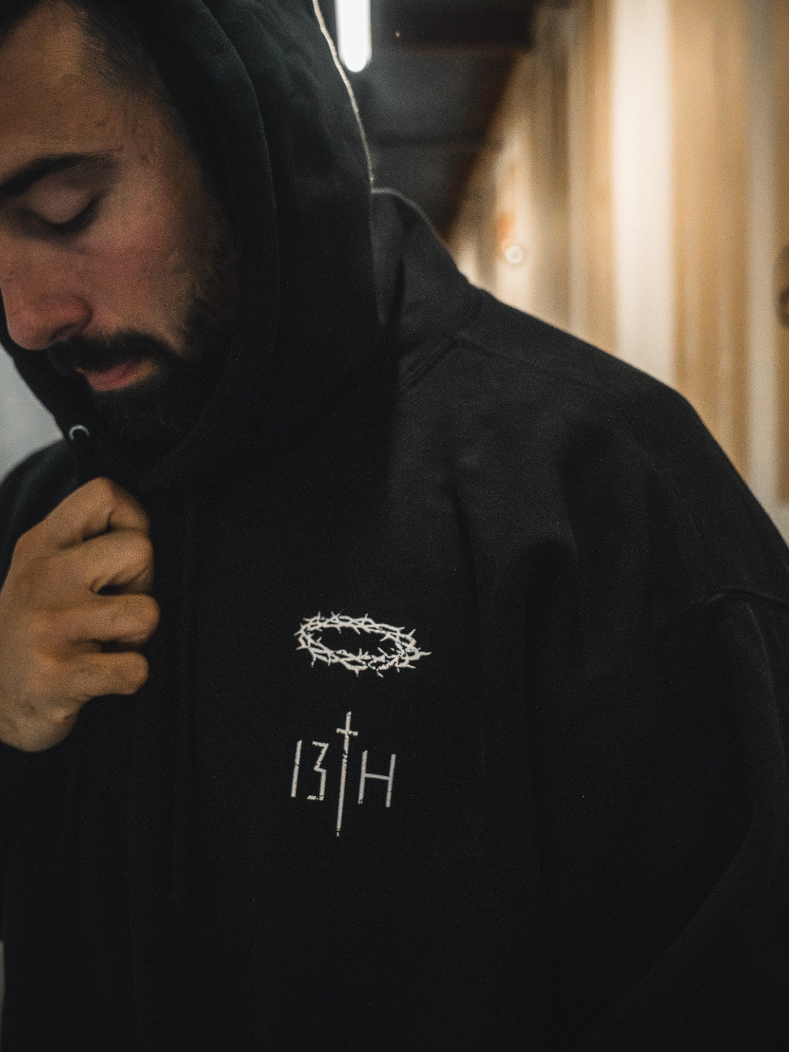 A person wears the black Jesus Saves Hoodie by 13th, hood up and holding the collar. The Christian hoodie shows a white crown of thorns above "13TH," with a sword as the letter T.