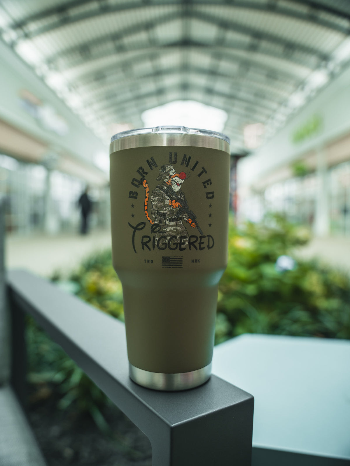 A 30oz tan Triggered Tumbler by Born United Private Label, featuring a coiled snake and “BORN UNITED TRIGGERED,” rests on a metal railing indoors with a glass ceiling and blurred plants in the background.
