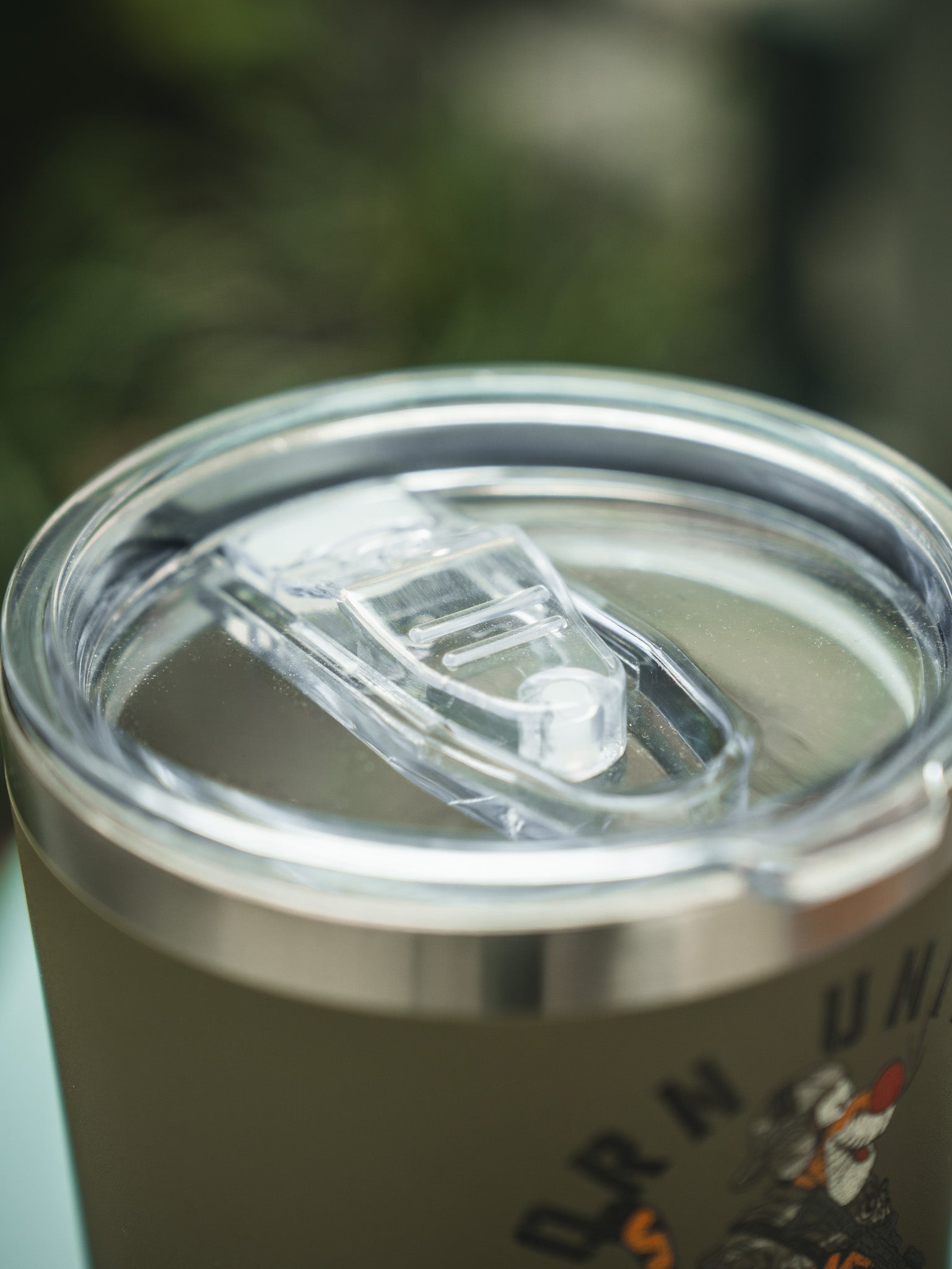 A close-up of the Born United Private Label Triggered Tumbler - 30oz, featuring stainless steel construction, a clear plastic lid with drinking spout, and partial graphic design on the side against blurred green foliage.