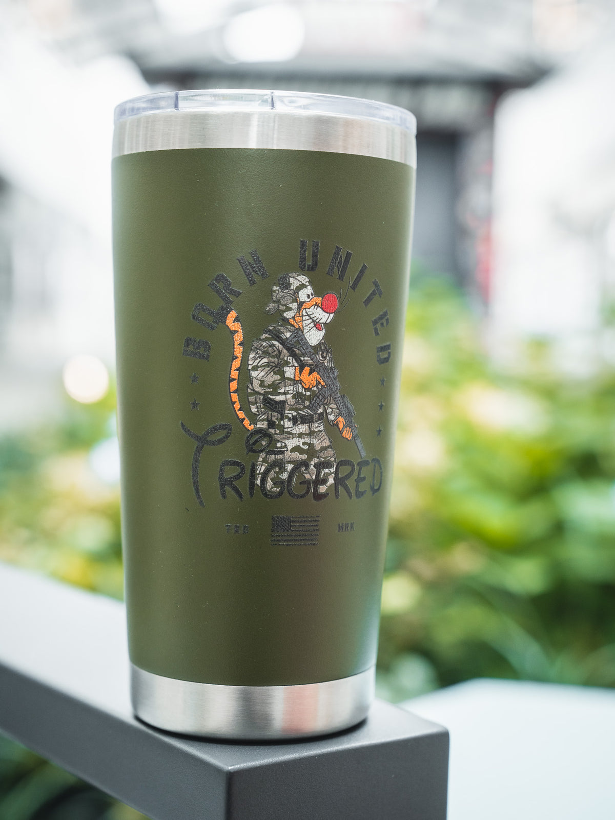 The Triggered Tumbler - 20oz by Born United Private Label is green with a metal rim, featuring a camo-clad baboon holding a rifle, "Born United Triggered" text above, an American flag below, and a blurred greenery background.